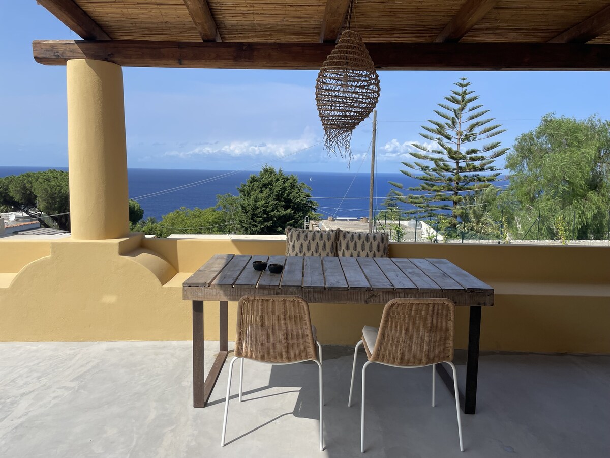 An outdoor terrace features a wooden dining table accompanied by two chairs, offering a 180° view of the sea and surrounding greenery. A woven pendant light hangs above the table, complementing the natural setting and fostering an inviting space for dining or relaxation.