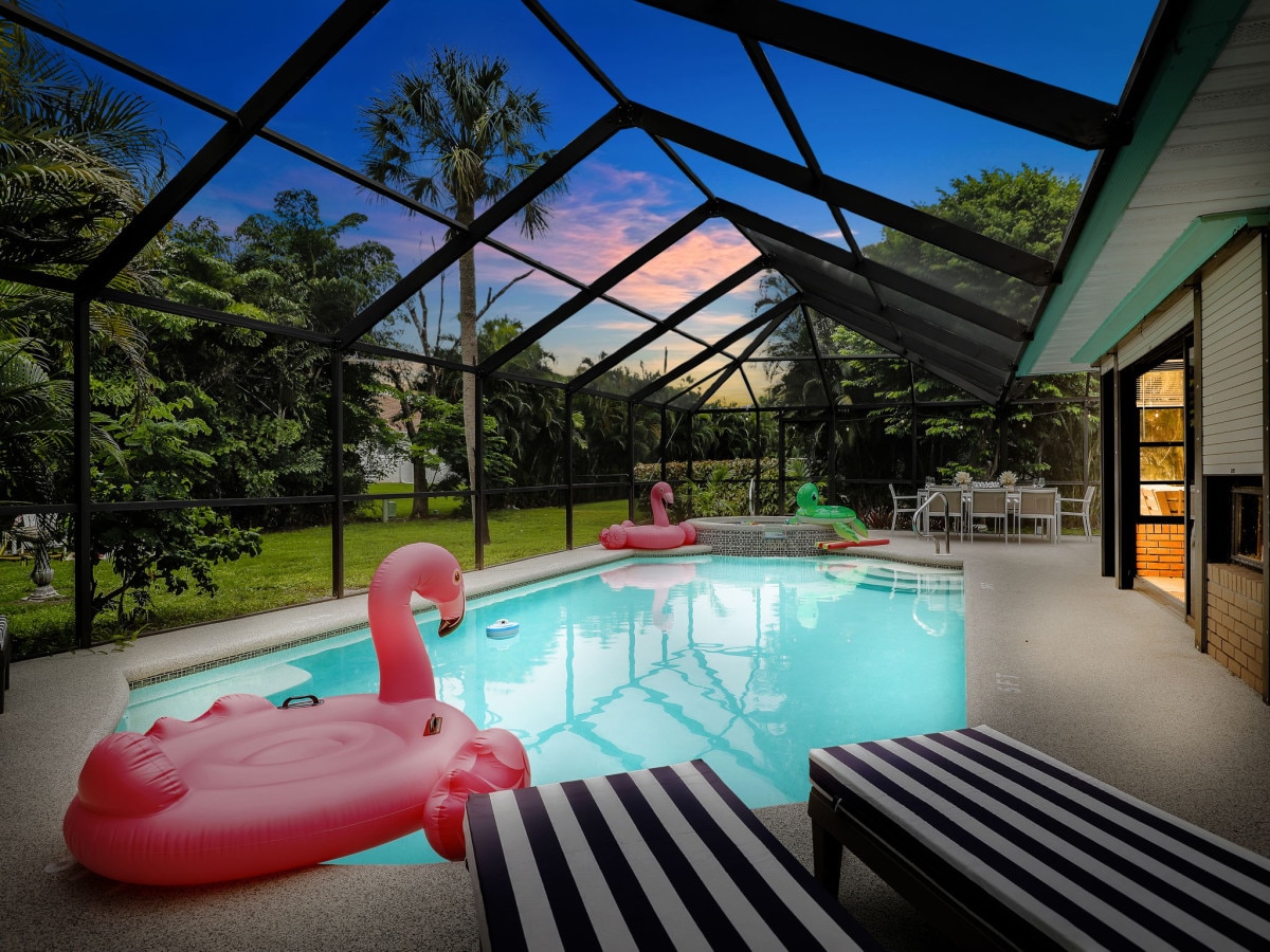 A heated pool is surrounded by a screened lanai, featuring two inflatable flamingo floats resting on the water. Lounge chairs with striped cushions are positioned beside the pool, offering a view of the well-maintained garden and evening sky.