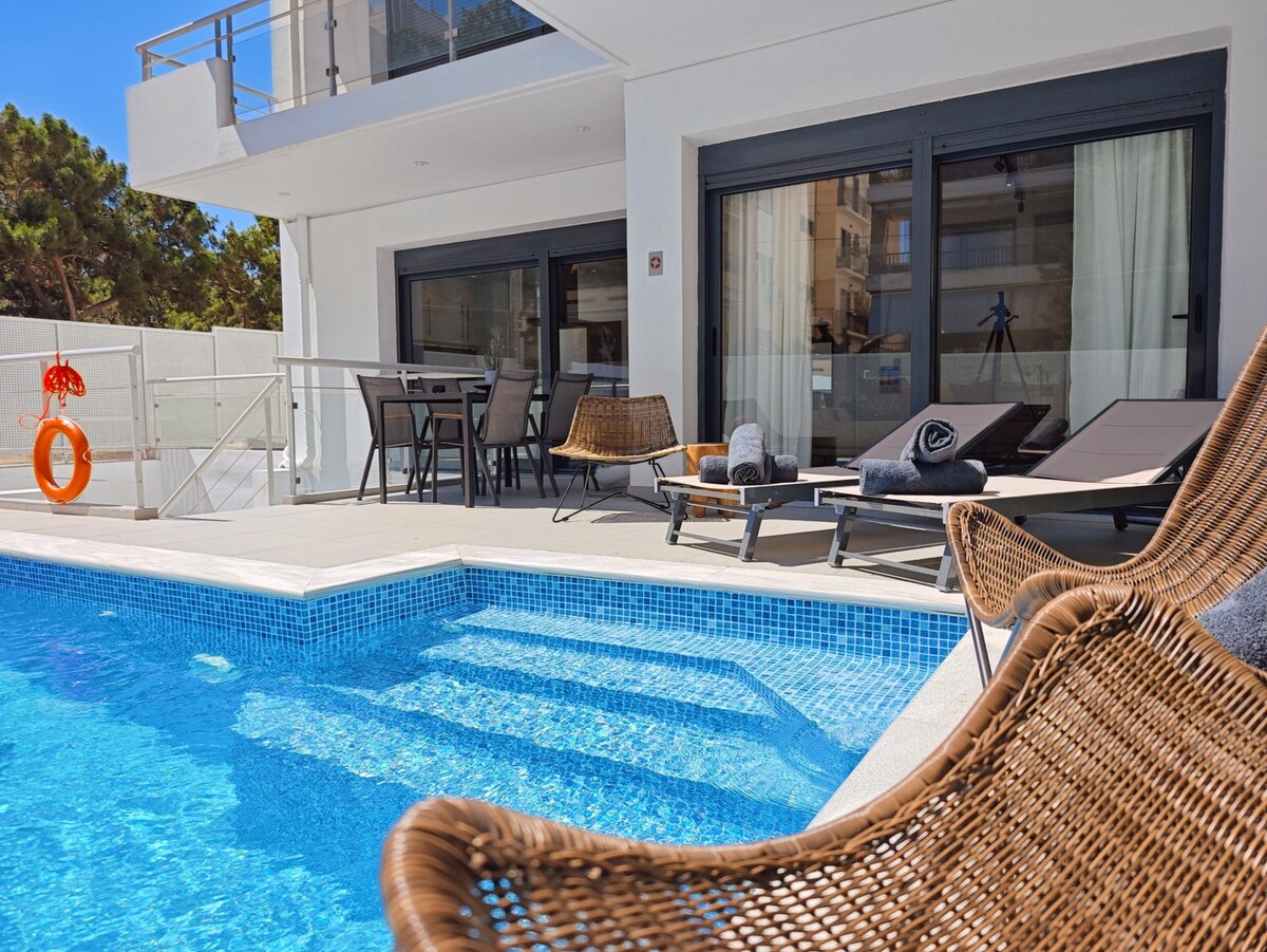 A private pool area is illuminated by sunlight, featuring a clear blue pool with steps leading down into the water. Modern loungers are positioned nearby, alongside a dining table set with chairs, framed by large glass doors that open into the living space.