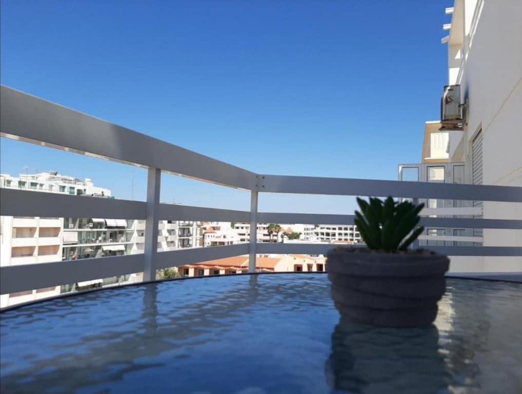 A balcony setting is depicted, featuring a glass table topped with a small potted plant. A clear blue sky is visible in the background, complementing the view of nearby buildings and rooftops.