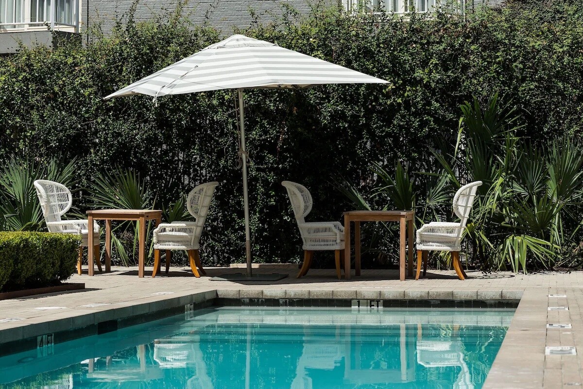 An outdoor swimming pool area is presented, featuring clear blue water surrounded by a stone deck. White umbrellas provide shade above several wicker chairs and small wooden tables, with lush greenery forming a natural backdrop.