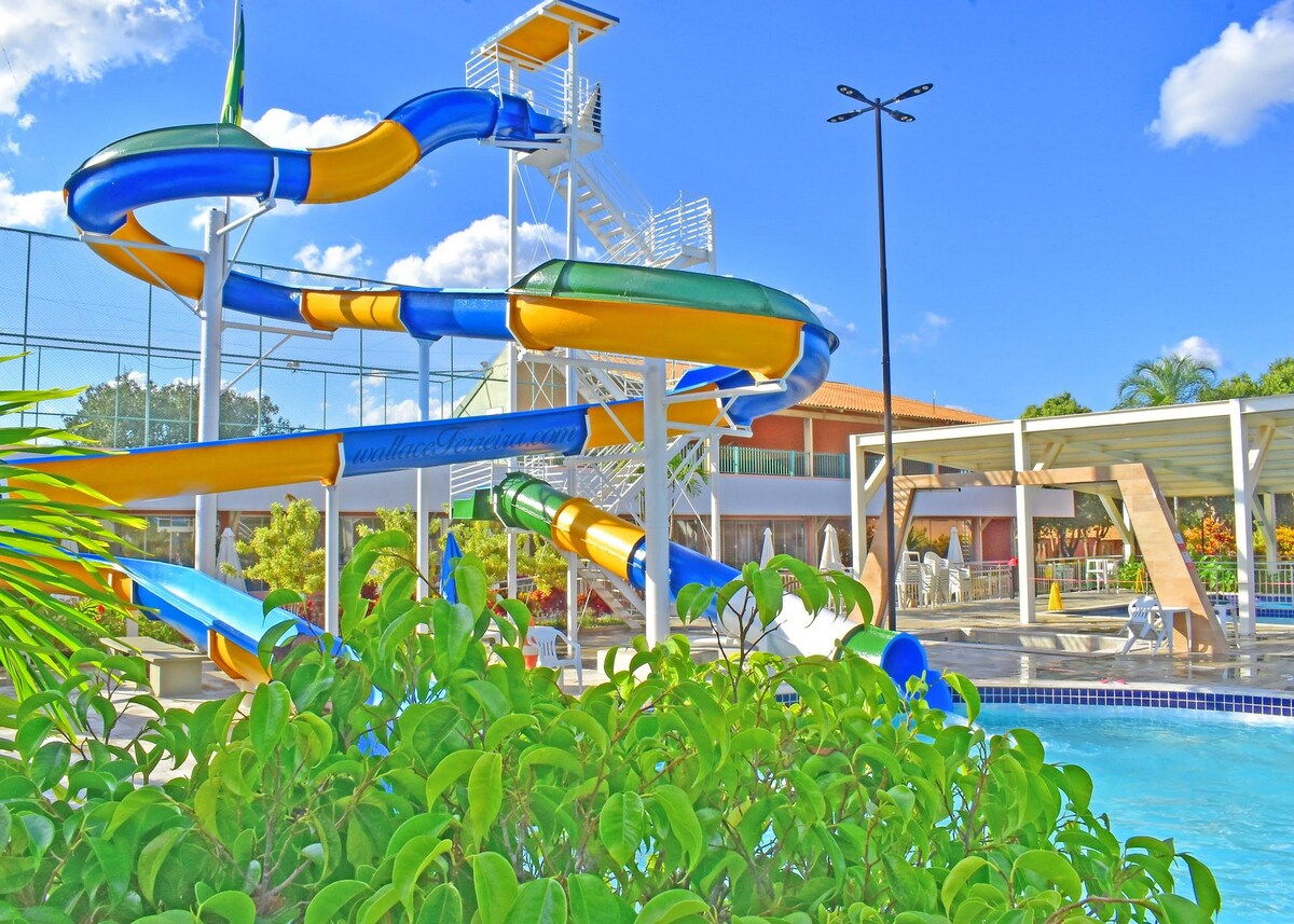 A vibrant water slide stands alongside a pool, surrounded by lush greenery. The slide features a colorful design with blue, yellow, and green sections. A modern structure is visible in the background, complementing the recreational atmosphere created by sunlit skies and inviting pool areas.