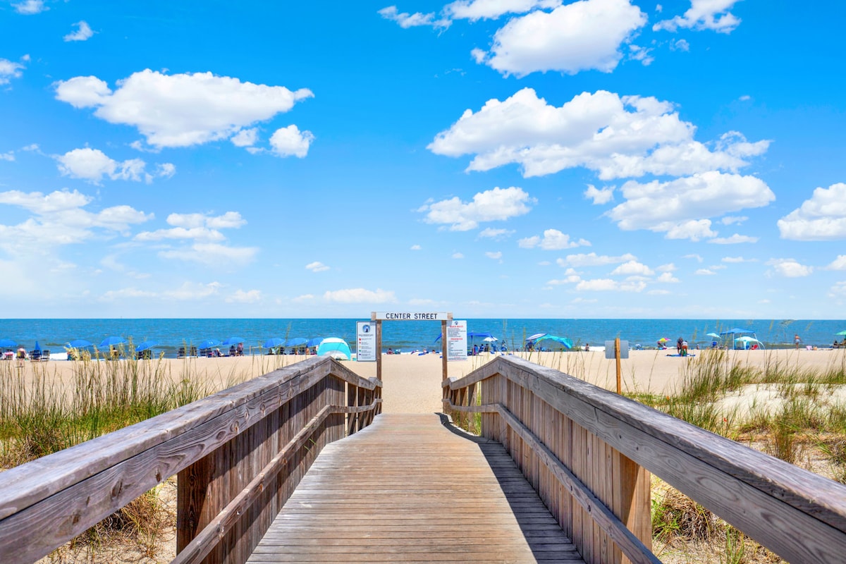 A wooden boardwalk stretches toward the beach, leading to a marked entry point. The sandy shoreline is visible, accompanied by umbrellas and beachgoers enjoying the sun. Grass lines the sides of the boardwalk, and the clear blue sky is dotted with fluffy clouds.