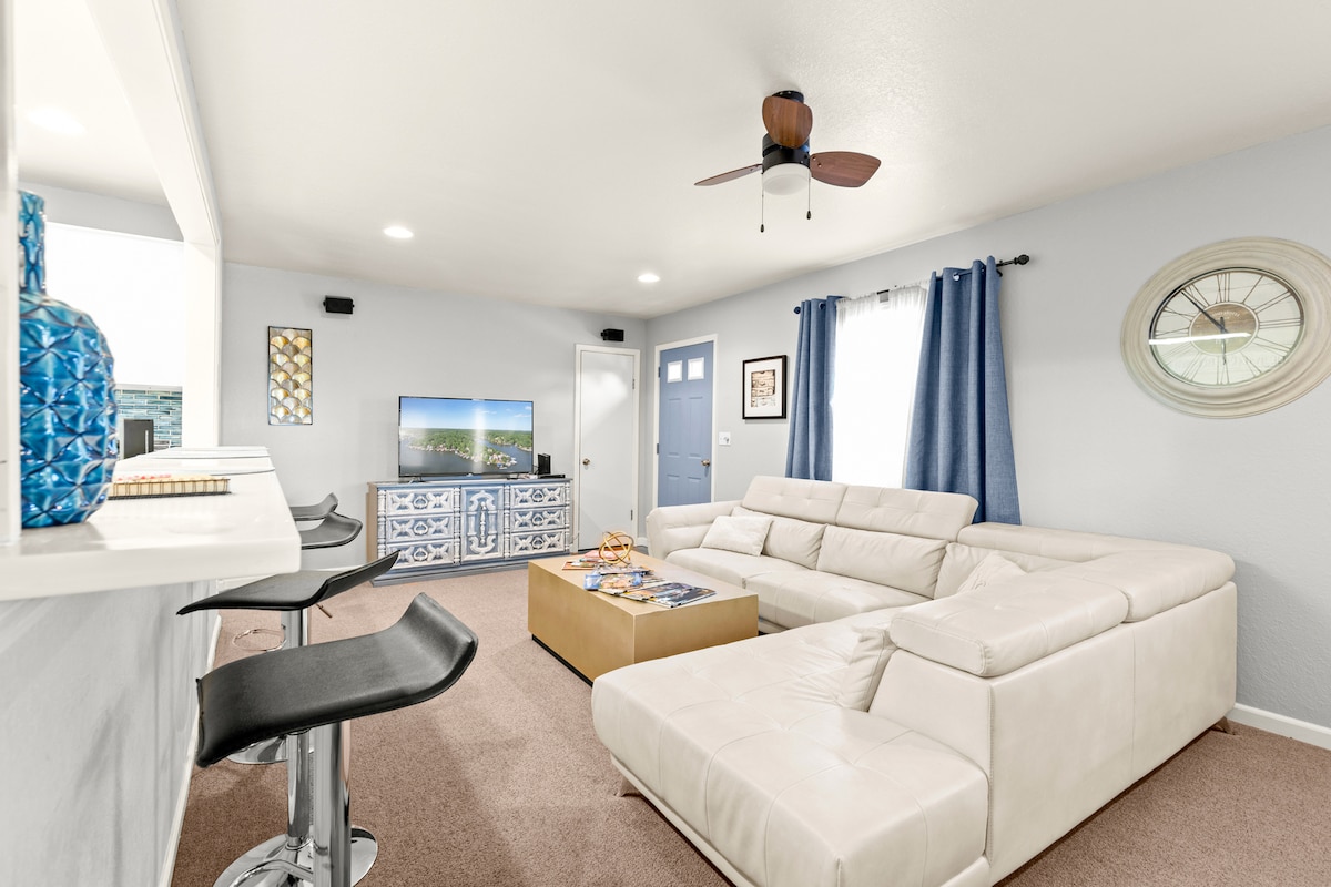 A light-filled living area features a sectional sofa, facing a media console with a television. A bar counter with seating is visible in the background. Decor includes a wall clock and decorative vases, complemented by window treatments that allow for natural light.
