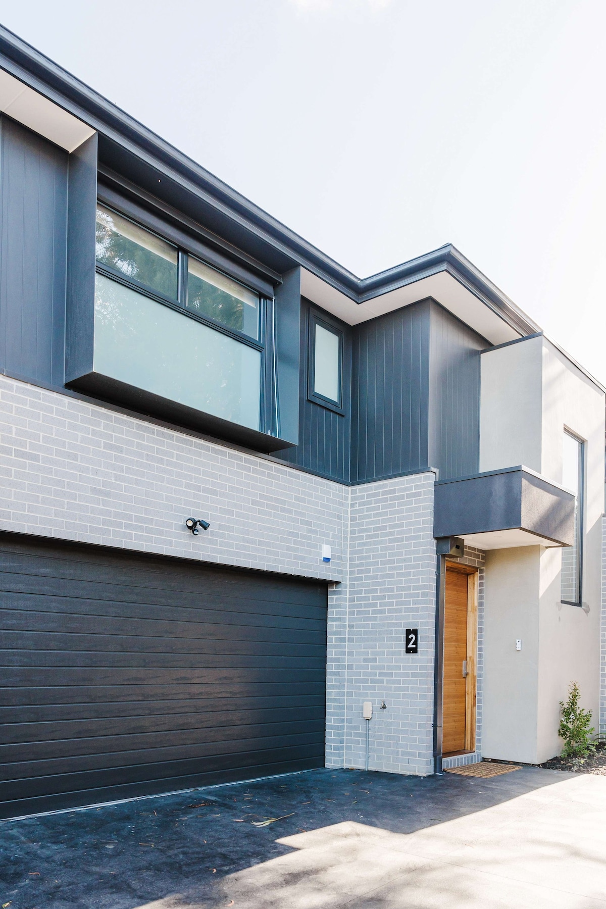 The modern townhouse exterior showcases a combination of grey brick and sleek paneling. A large garage door sits to the left, while a wooden entrance door framed by a minimalist overhang is visible on the right. The large windows invite natural light into the upper level.