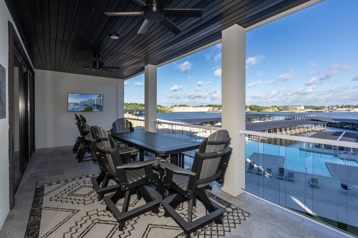 A spacious outdoor patio is furnished with a large dining table surrounded by comfortable chairs. An outdoor television is mounted on the wall, and stunning views of marina docks and a pool are visible beyond the balcony railing. The area is brightened by sunlight and blue skies.