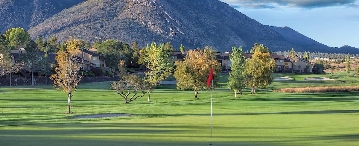 A scenic golf course is set against the backdrop of majestic mountains, featuring lush green fairways and strategically placed trees. A flag marks the location of a putting green, highlighting the serene outdoor environment ideal for leisure and recreation.