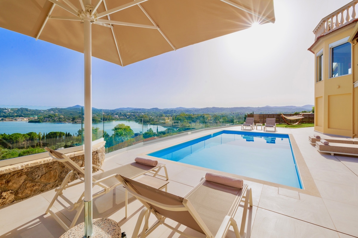 A private swimming pool is framed by a spacious patio area, complete with high-quality sunbeds and a large umbrella for shade. The pool area offers panoramic views of the surrounding landscape and glistening sea, illuminated by the bright sun.