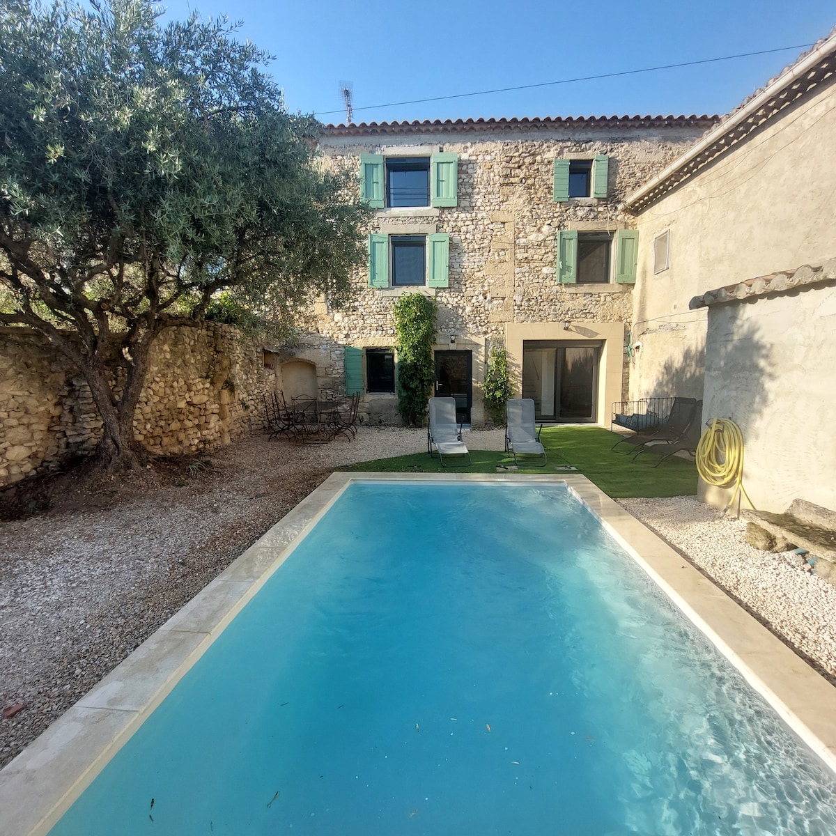 A serene courtyard features a swimming pool surrounded by lush greenery and a stone farmhouse. Sun loungers are positioned near the pool, while dining tables are visible in the background, complemented by olive trees and a gravel path.