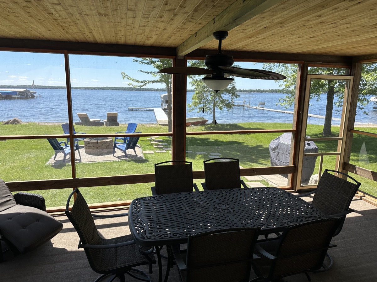 A screened-in porch is shown with a large outdoor dining table surrounded by several chairs. The view reveals a grassy area leading to a tranquil lake with seating areas and a dock beyond. Natural light filters through large screens, enhancing the spacious feel.
