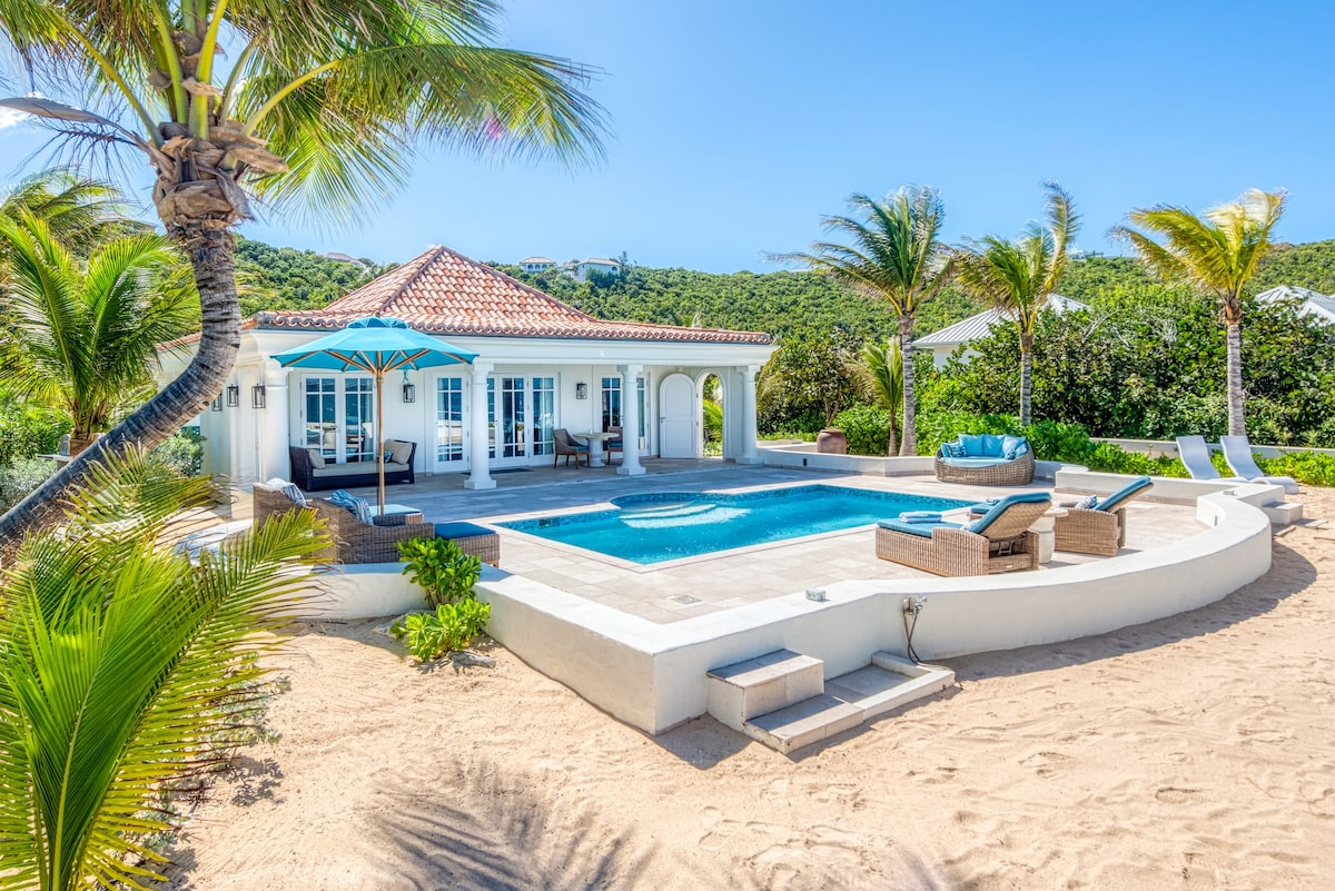An airy villa is set against a backdrop of tropical greenery and clear blue skies. A serene pool reflects sunlight, while comfortable lounge chairs invite relaxation. In the distance, palm trees frame the scene, creating a peaceful beachfront atmosphere.