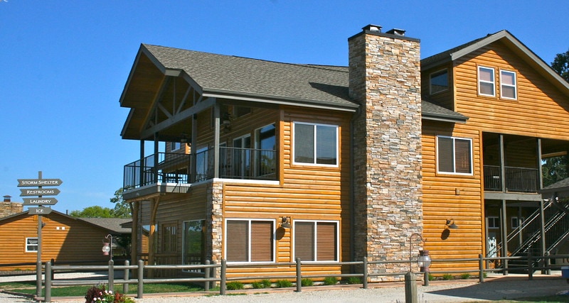 The exterior of a large wooden building features a stone chimney and wooden accents. A spacious balcony is visible on the upper level, while multiple windows reflect natural light. Signposts indicating nearby amenities are positioned in the foreground, contributing to the welcoming atmosphere.