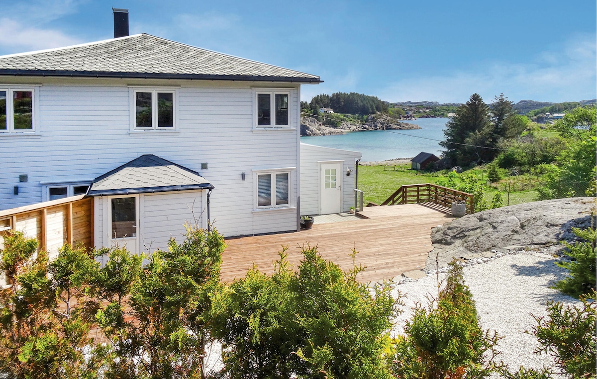The exterior of the holiday home is displayed, featuring a light-colored façade and a spacious wooden terrace. A view of the tranquil water and green landscape is visible in the background, complemented by nearby natural elements.