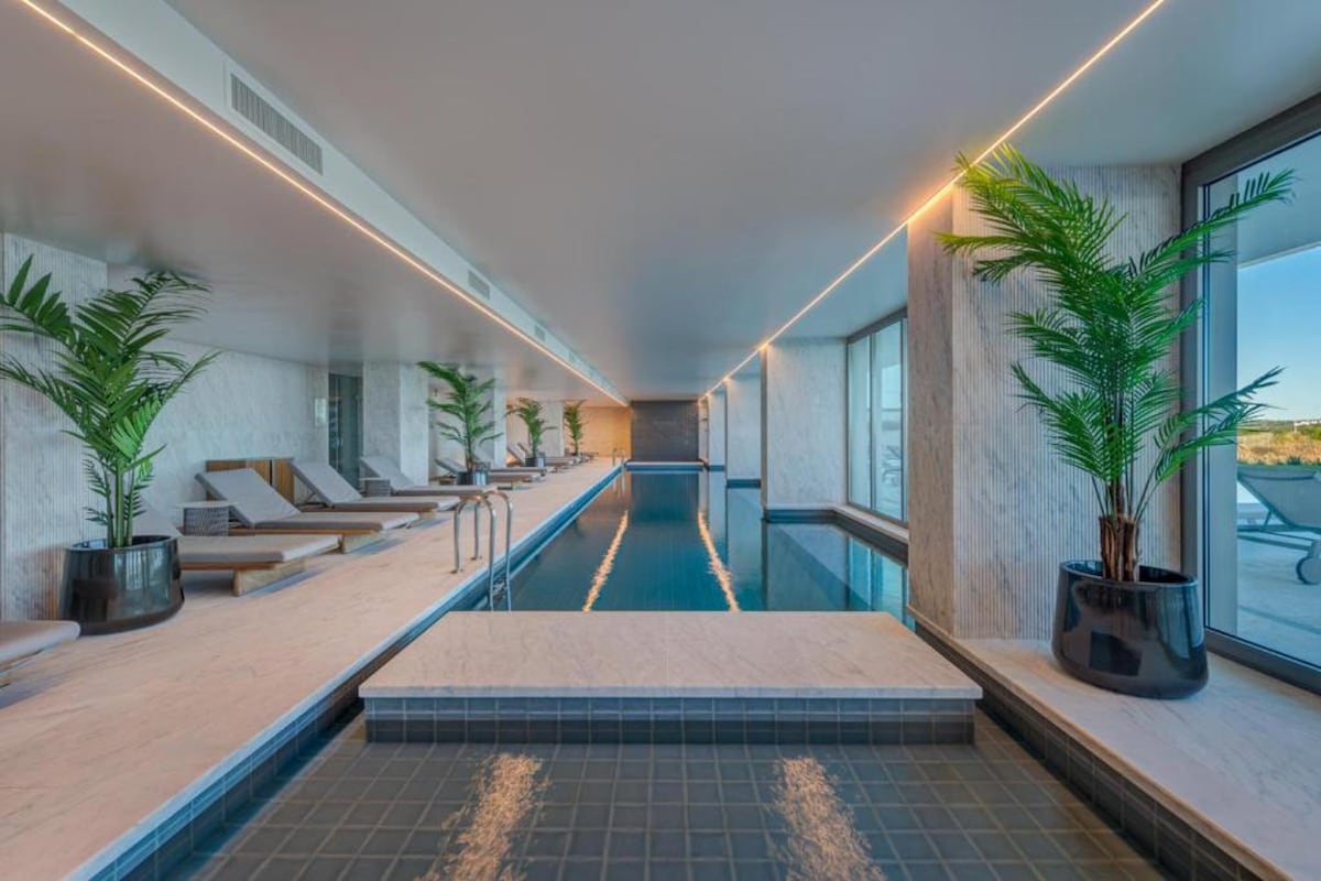 An indoor swimming pool is framed by sleek marble walls and lined with comfortable lounge chairs. Potted plants add a touch of greenery, while large windows allow natural light to illuminate the tranquil water, creating a serene environment for relaxation.