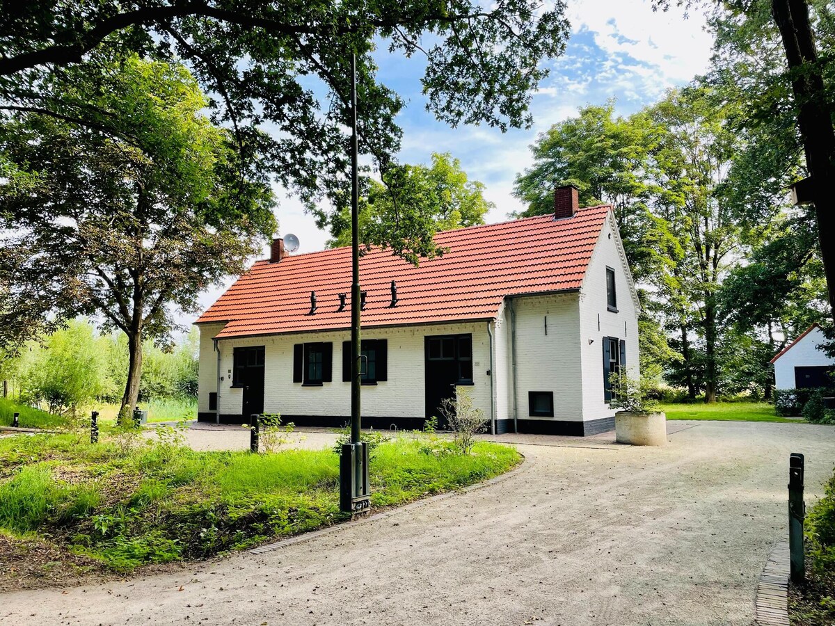 The holiday home is set amidst lush greenery, featuring a traditional exterior with white walls and a red-tiled roof. The building is partially surrounded by trees, with a gravel pathway leading up to it, offering a serene entrance.