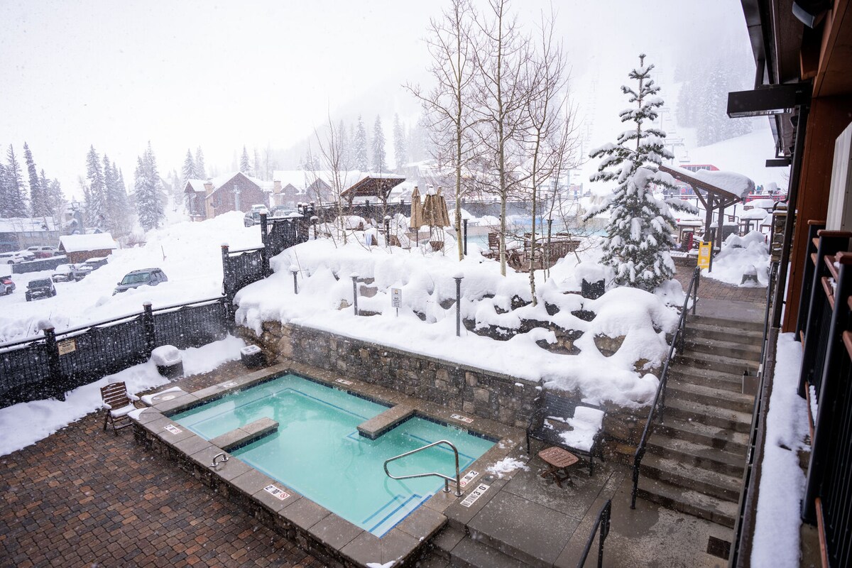 A secluded outdoor hot tub is surrounded by a snow-covered landscape, showcasing a serene atmosphere. Steps lead down to the tub area, with snow-draped trees and a few lounge chairs visible. Light snowfall adds to the winter scene, enhancing the tranquil environment.