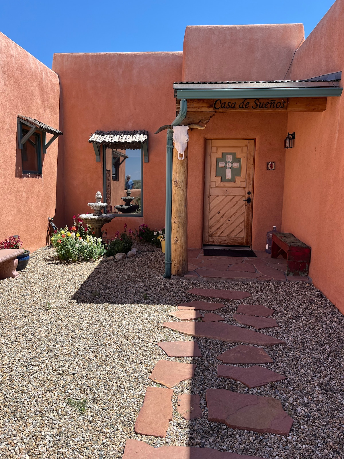 The entrance to Casa de Suenos is framed by adobe-style walls in warm terracotta tones, featuring a decorative wooden door with a unique design. A charming garden area showcases colorful flowers, and a stone pathway leads up to the door, creating an inviting approach to the home.