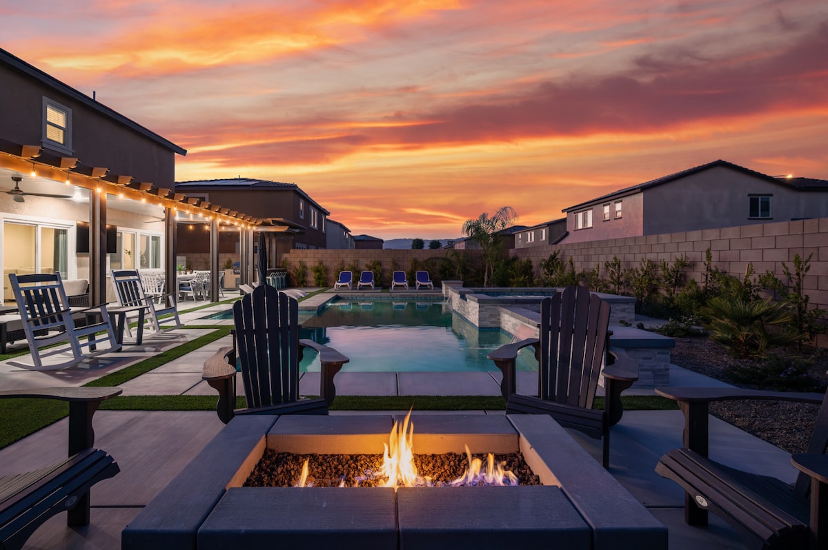 The outdoor area features a heated pool surrounded by lounge chairs and a fire pit with a flame at its center. A patio provides seating under a wooden pergola, while vibrant skies showcase hues of orange and pink at sunset.