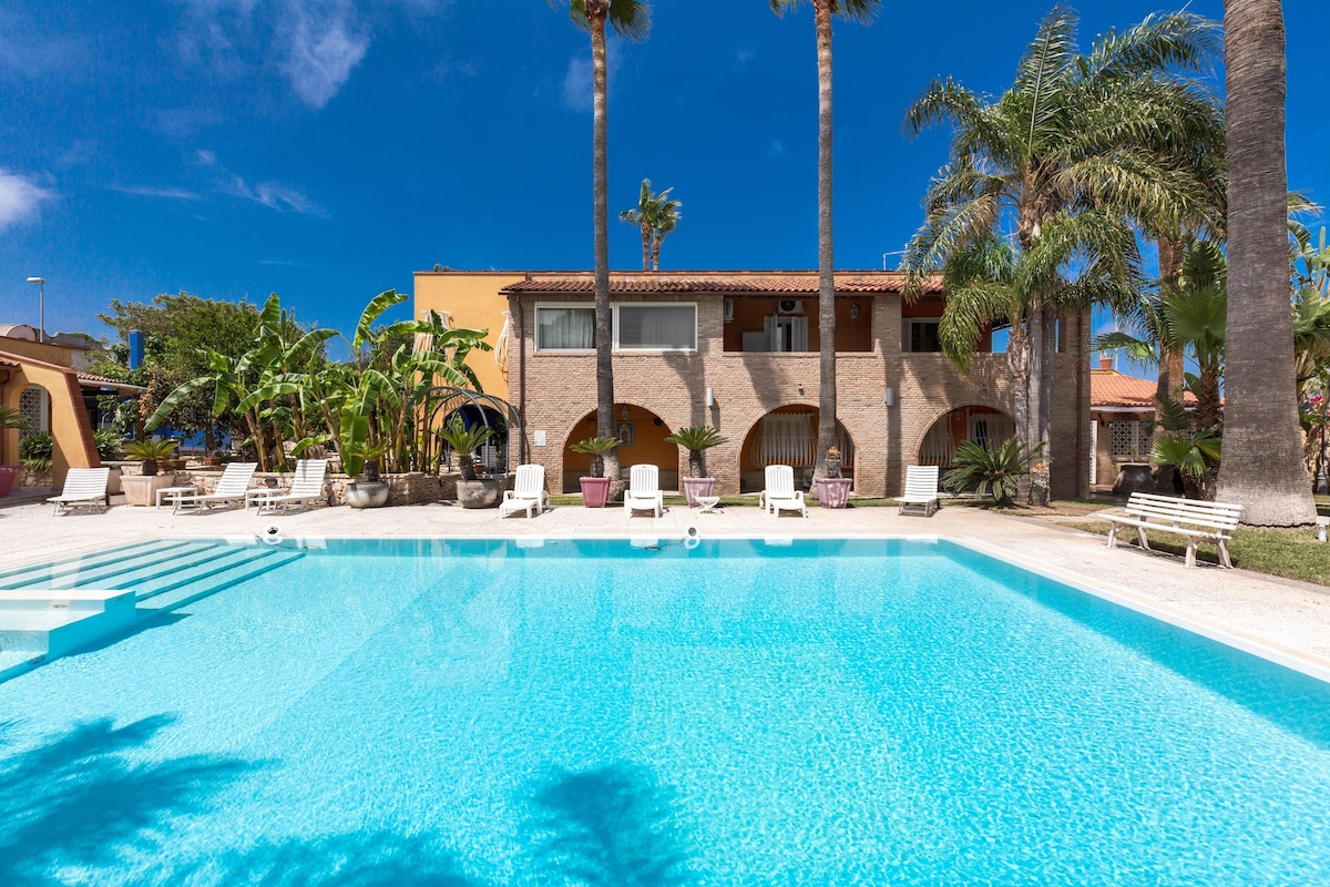 A large outdoor pool is surrounded by a spacious deck with white lounge chairs. Towering palm trees can be seen alongside the vibrant greenery, while a two-story villa in warm tones stands in the background under a clear blue sky.