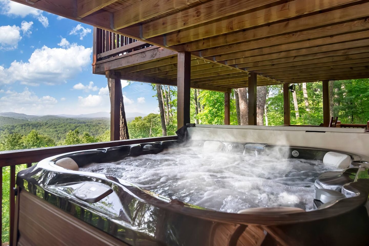A spacious hot tub is positioned on the deck, surrounded by natural greenery. Gentle steam rises from the water, hinting at warmth. The view in the background showcases rolling hills and a clear blue sky, adding to the tranquil outdoor setting.