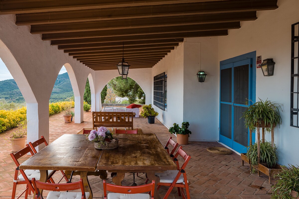 An outdoor terrace is featured, framed by arched columns and a wooden roof. A rustic wooden table is surrounded by six chairs, and potted plants add a touch of greenery. Scenic views of mountains and gardens are visible in the background.
