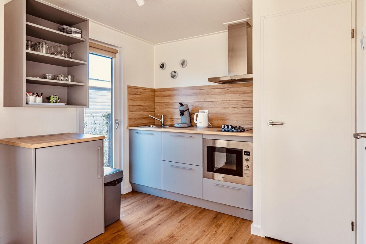 A compact kitchen area features modern cabinetry and a wooden countertop. Essential appliances, including a microwave and coffee maker, are visible. A window allows natural light to enter, enhancing the simplicity of the design. A small table is positioned adjacent to the counter.