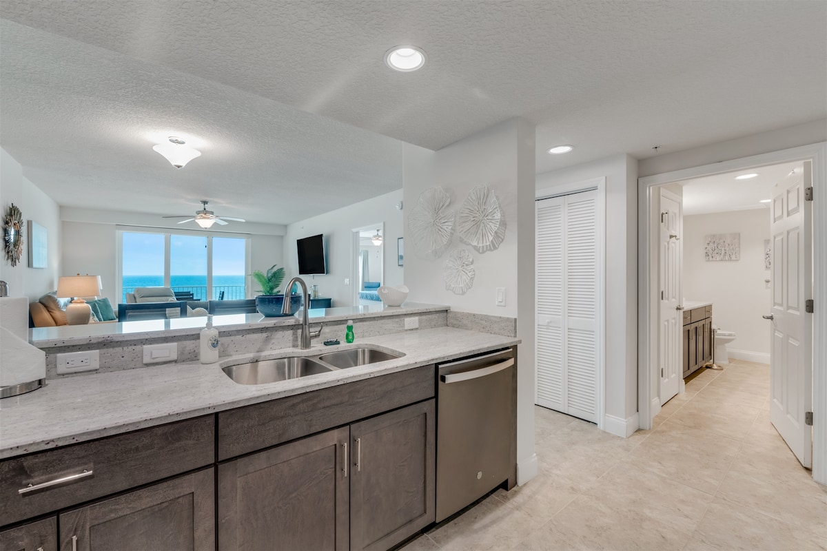A modern kitchen space is showcased, featuring sleek countertops and stainless steel appliances. The open layout connects seamlessly to the living area, where expansive windows offer views of the ocean. Simple decor elements add a touch of elegance to the environment.