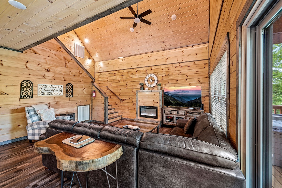 The living room features a spacious seating area with a large sectional sofa and a wood coffee table. Pinewood walls add warmth, while a flat-screen TV and a fireplace enhance the space. A staircase leads to the upper level, and large windows provide natural light.