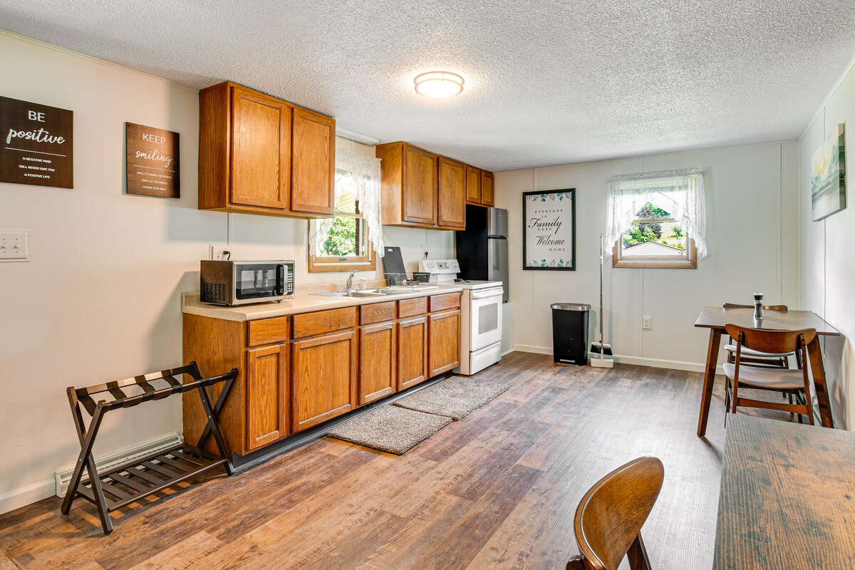 A well-equipped kitchen is complemented by wooden cabinets and essential appliances including a microwave and stove. A dining table with chairs occupies one corner, while decorative wall art adds a personal touch. Natural light enters through a window adorned with lace curtains.