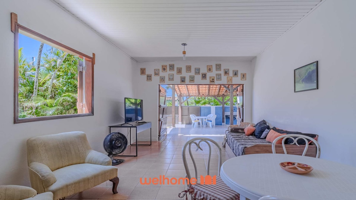 A bright living area is illuminated by natural light streaming through a large window. A comfortable seating arrangement includes a beige armchair and a sofa adorned with cushions. A circular dining table sits center, accompanied by a TV mounted on the wall. Greenery is visible outside.