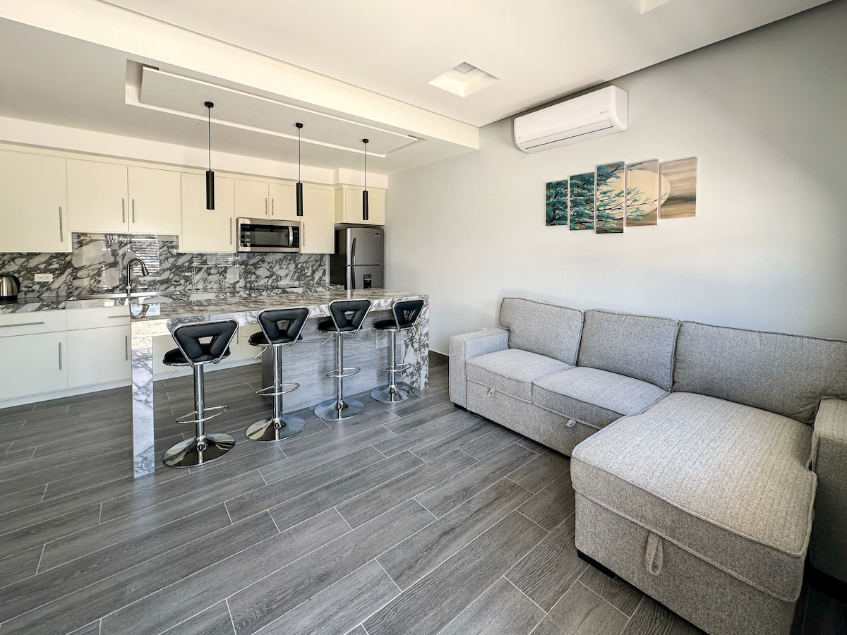 A modern kitchen area features sleek cabinetry and marble countertops. Four bar stools are positioned at the countertop. A spacious sectional sofa sits comfortably in the living area, complemented by neutral tones and natural light entering from nearby windows.
