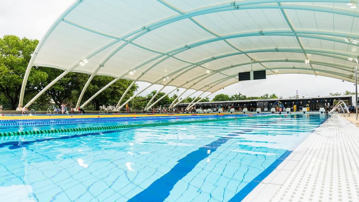 A spacious outdoor pool is sheltered under an expansive canopy, featuring clear blue water and marked lanes. Surrounding amenities such as seating areas and shaded spots are visible in the background, inviting relaxation. Lush greenery is present, enhancing the tranquil environment.