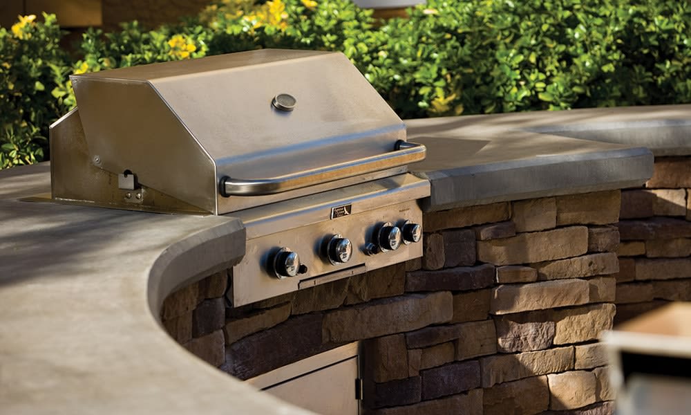 A stainless steel barbecue grill is integrated into a curved stone countertop, surrounded by green shrubs. The grill features three knobs and a lid that is slightly ajar, emphasizing its ready-to-use condition in the outdoor dining area.
