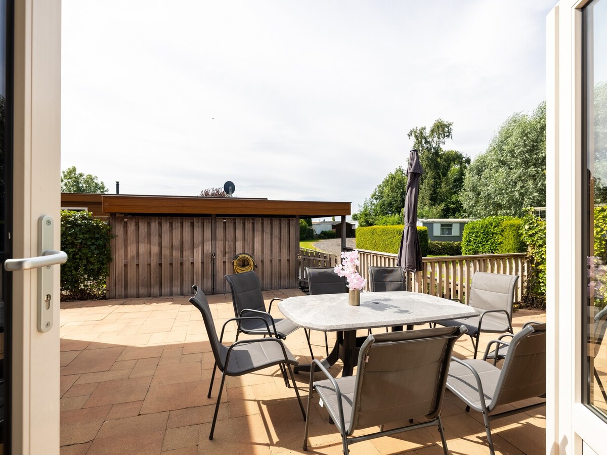 A private terrace is visible, featuring a round table surrounded by six chairs. A large umbrella provides shade, while a wooden fence adds privacy. Lush greenery surrounds the area, contributing to a serene outdoor setting ideal for relaxation.