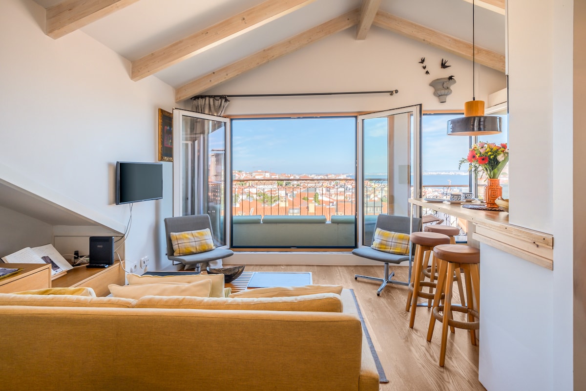 A modern living space features large glass doors that open to a balcony, allowing for ample natural light. Two armchairs and a sofa are arranged to provide a comfortable seating area. A bar with stools is situated next to the kitchen area, enhancing the open layout.