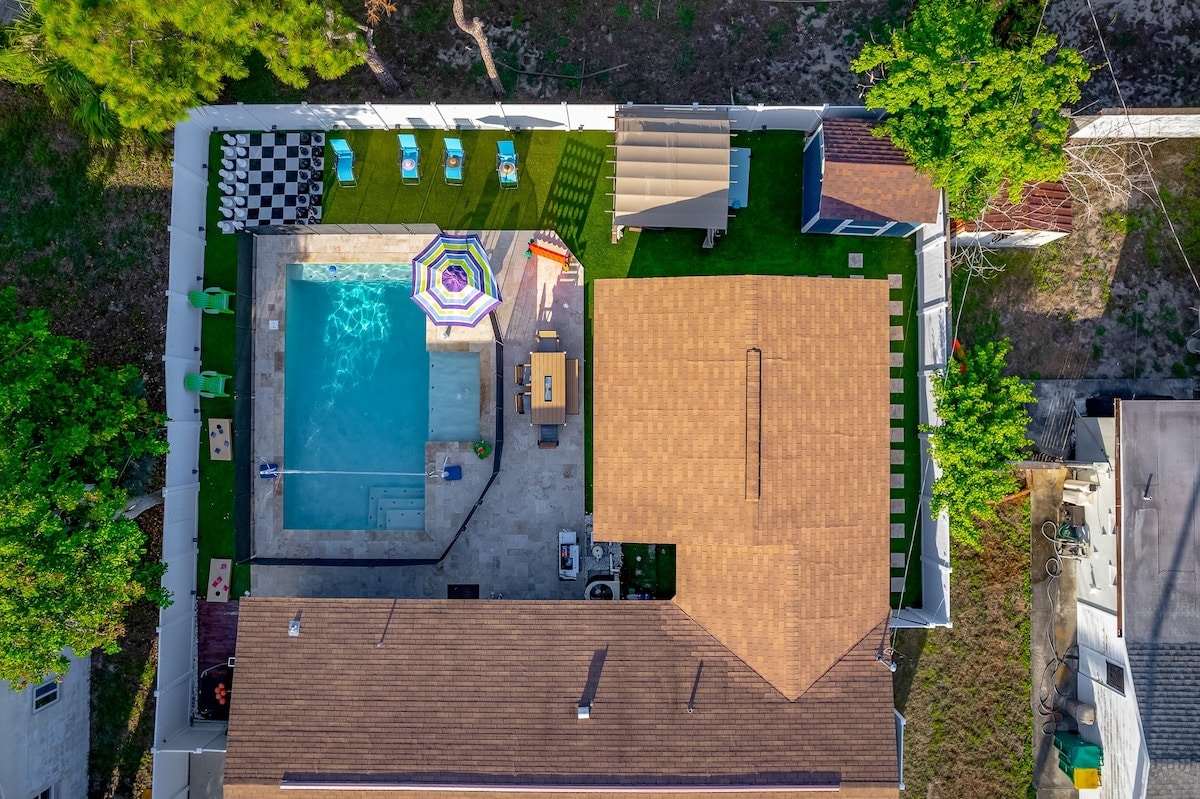 An aerial view of a vibrant outdoor space features a heated pool surrounded by lounge chairs. A shaded area provides a space for dining, while colorful games are placed around the manicured lawn. The home extends cozy living to the outdoors, inviting relaxation and play.