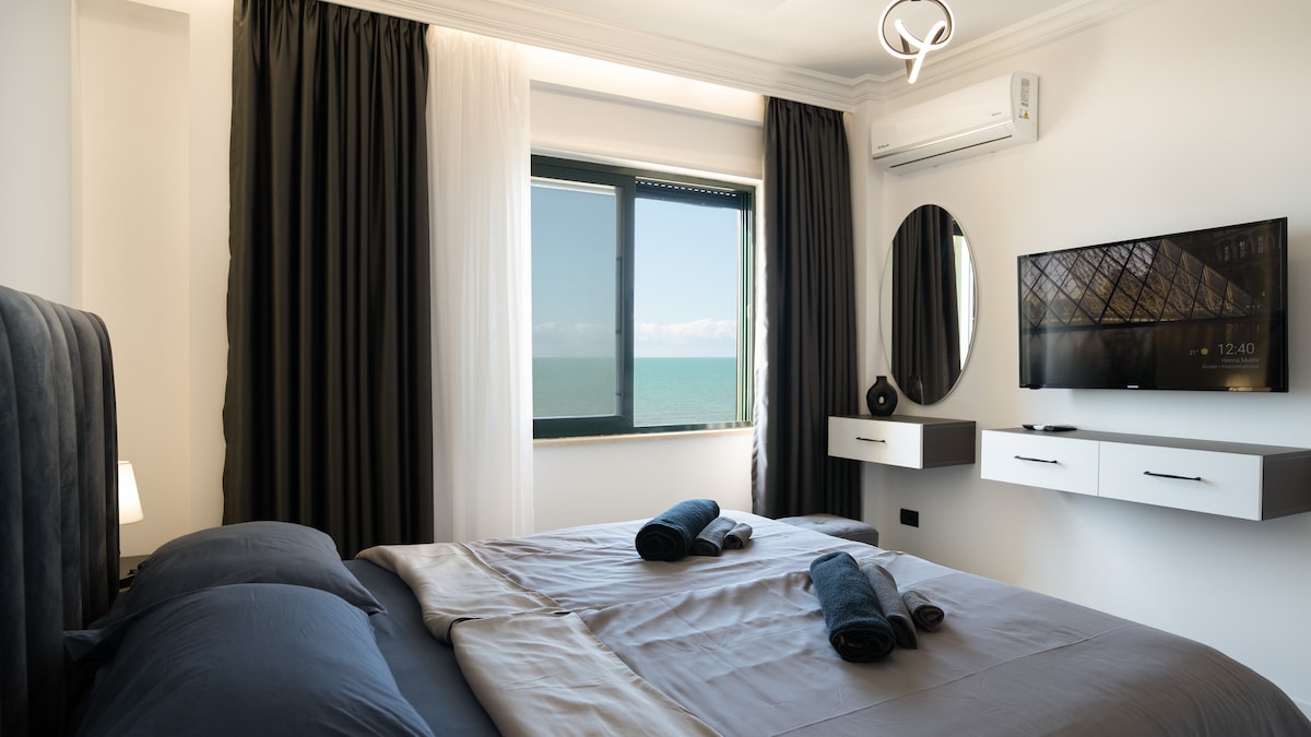 The bedroom features a spacious bed adorned with neatly arranged towels. A wall-mounted flat-screen TV is positioned opposite a large window that frames a view of the ocean. Dark curtains contrast with light drapes, complementing the contemporary decor.