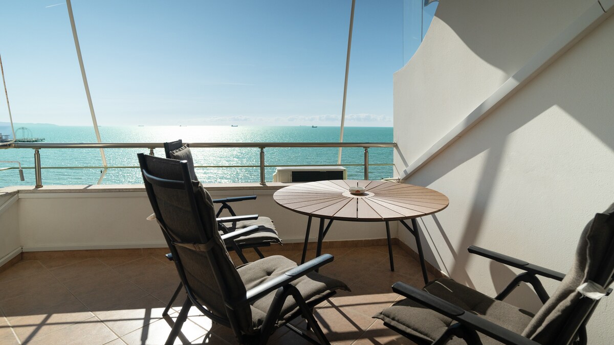 A spacious terrace features a round table and two comfortable chairs, inviting relaxation. Bright natural light illuminates the area, highlighting stunning sea views that stretch across the horizon, with gentle waves glistening in the sunlight.