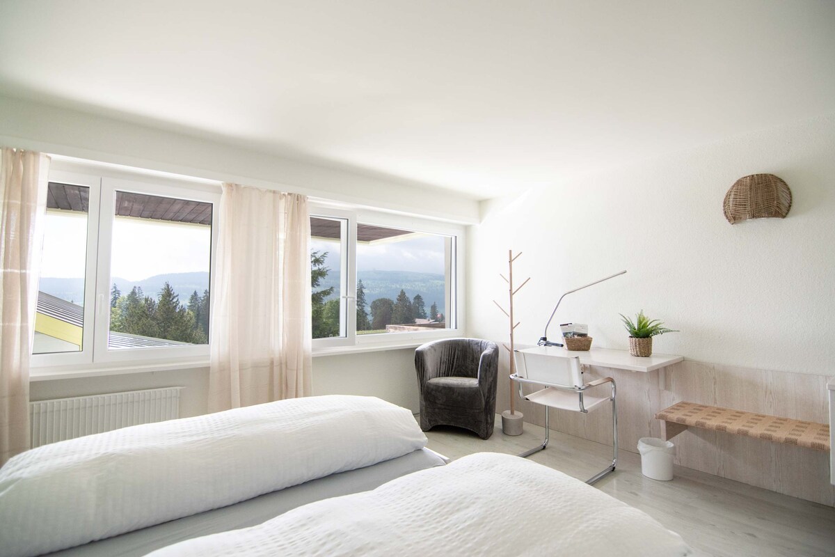 This bright room features twin beds with white linens and a desk. Large windows provide a view of the surrounding nature. A comfortable chair is placed in the corner, and a small plant sits on the desk, creating a serene and functional space.