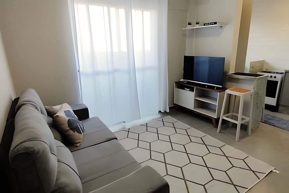 A cozy living area is shown with a grey sofa facing a mounted flat-screen TV. Light filters through sheer curtains, creating a bright ambiance. A light-colored geometric rug lies on the floor beneath a small white stool and a minimalist shelf displaying decorative items.