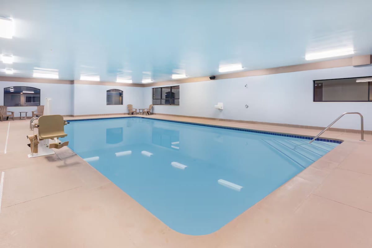 An indoor swimming pool is showcased, featuring a clear blue water surface and a gently sloped entry. The spacious area is illuminated with bright ceiling lights, and a chair lift is positioned for accessibility. Large windows provide a view of the exterior while bathing the room in natural light.