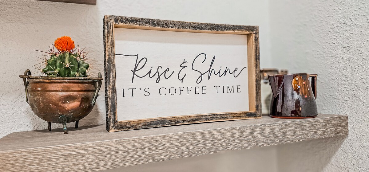 A wooden shelf is showcased with a framed sign reading 'Rise & Shine, It's Coffee Time.' An orange-flowered cactus sits in a metallic pot beside a dark ceramic coffee pot, creating a charming vignette.