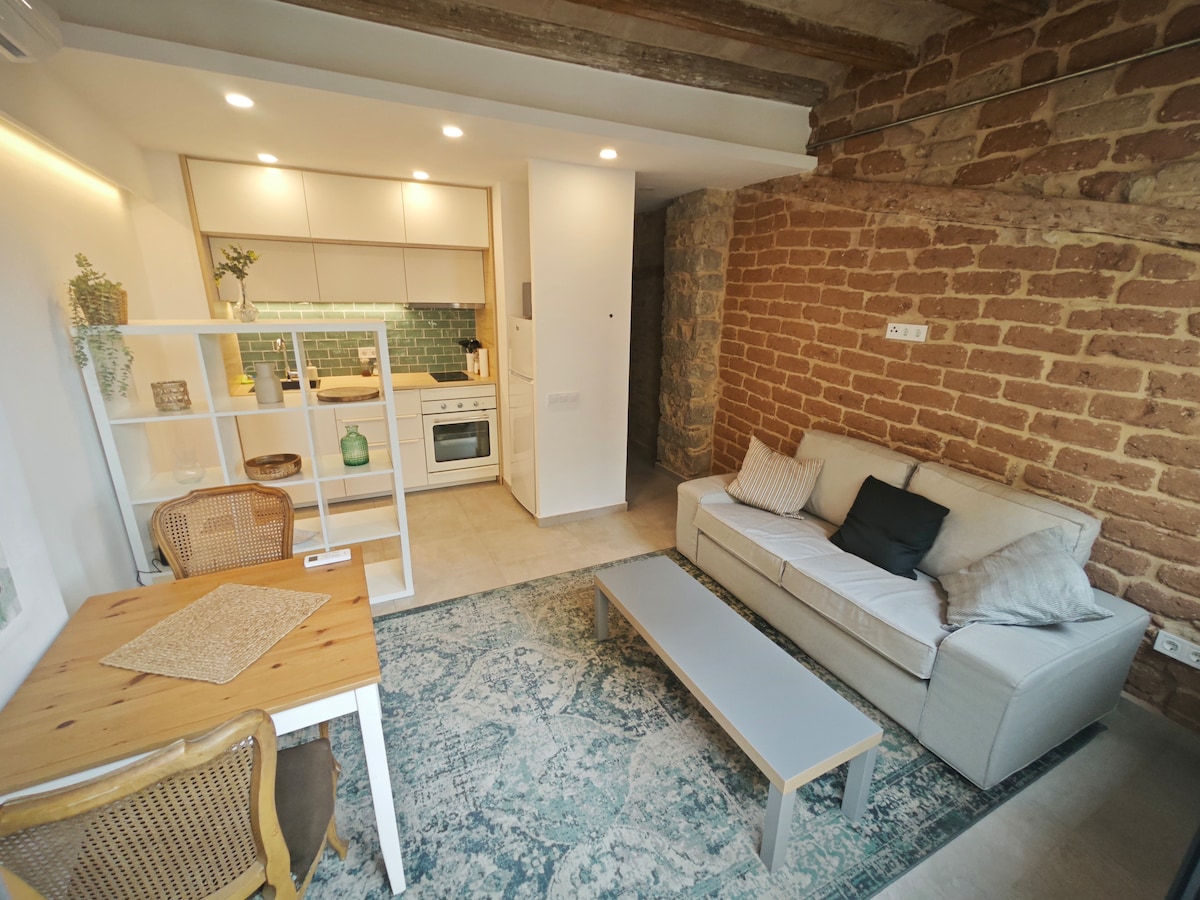 A modern living area features exposed brick walls and a comfortable seating arrangement. A light wood dining table is accompanied by a stylish chair. The kitchen is equipped with contemporary appliances, and soft lighting creates an inviting ambiance throughout the space.