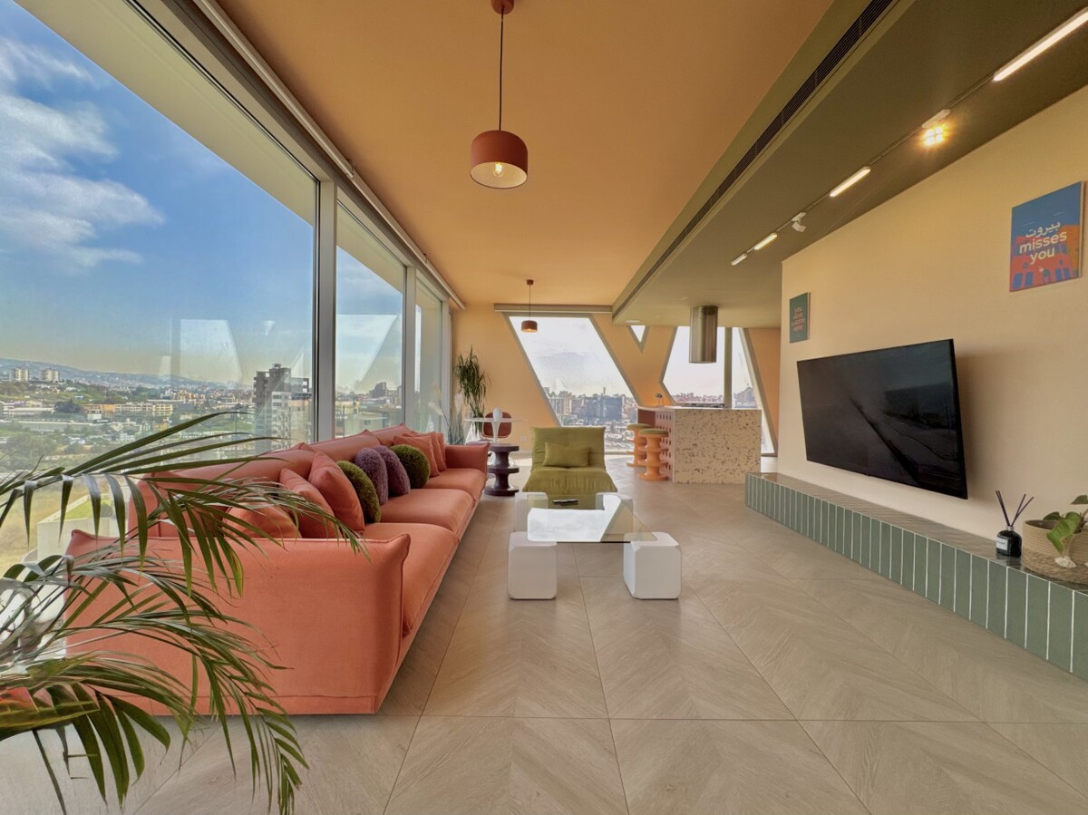The spacious living area features large floor-to-ceiling windows, offering panoramic views of the city. A modern orange sofa is complemented by colorful cushions, while a sleek white coffee table is positioned on a light tiled floor. A flat-screen TV is mounted on the wall, and greenery adds a touch of nature.