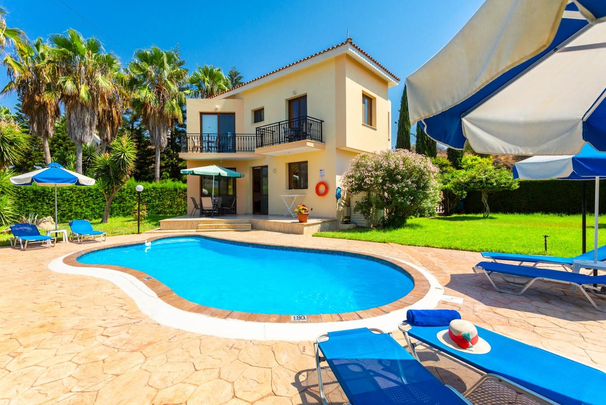 A spacious villa features a private pool surrounded by sun loungers and umbrellas, nestled in a lush garden. The two-story structure showcases a balcony with access from a set of double doors. A vibrant poolside area includes a small table set for outdoor dining.