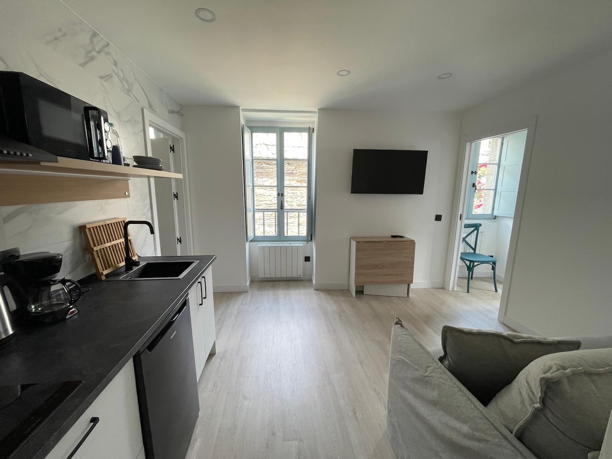 An open living space features a modern kitchen area with sleek black countertops and a coffee maker. A comfortable sofa faces a wall-mounted TV, while large windows allow natural light to fill the room. A wooden cabinet and a green chair add to the decor.