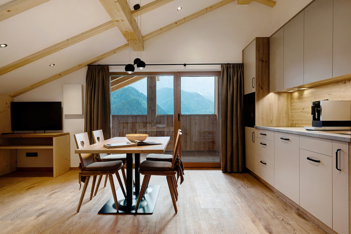 A bright and airy interior is presented, featuring a dining area with a central table surrounded by four chairs. Large windows offer views of the surrounding mountains, while a modern kitchen with stylish cabinetry is visible along one wall. Wooden beams create a spacious feel.