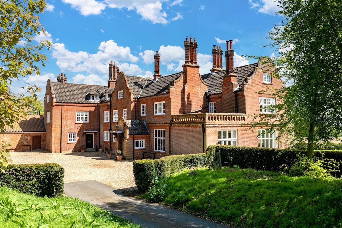 An elegant red-brick building showcases classic architecture with multiple gabled roofs and prominent chimneys. Surrounding greenery softens the view, while a gravel pathway leads to the entrance, inviting guests into a spacious estate set against a backdrop of blue sky and scattered clouds.