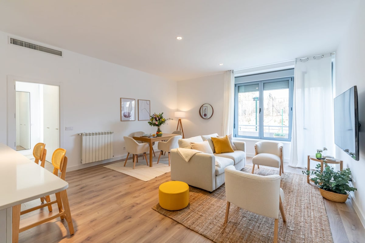 A bright living-dining area features a comfortable sofa, two armchairs, and a small round table with four chairs. Large windows allow natural light to fill the space, while decorative elements like a patterned rug and indoor plants add warmth. A wall-mounted television is positioned for easy viewing.