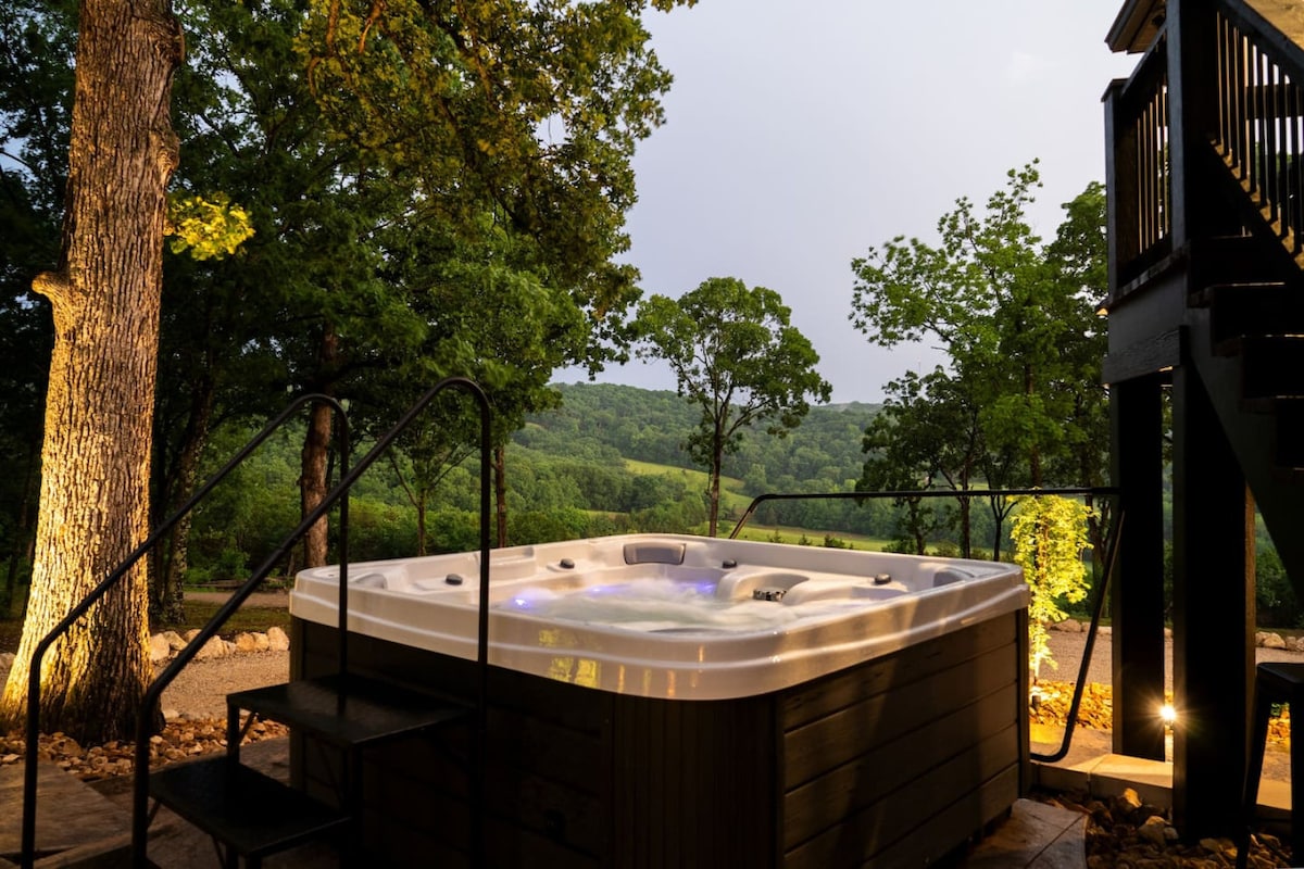 A hot tub is situated on a wooden deck, surrounded by lush greenery and trees. Soft lighting enhances the ambiance, while scenic views of the landscape are visible in the background.